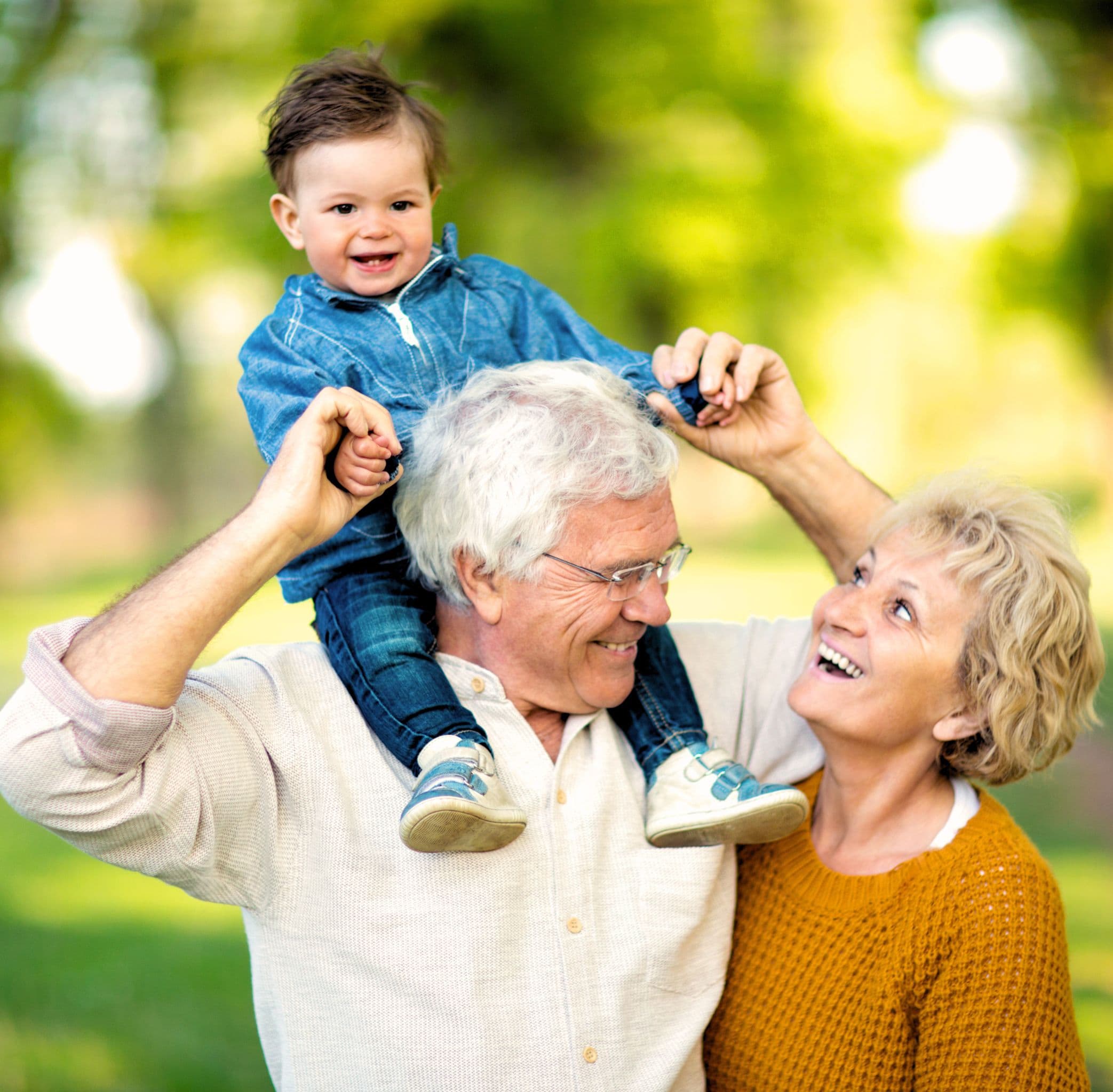 Grandparents outdoors in the green, their grandchild laughing on grandpa’s shoulders